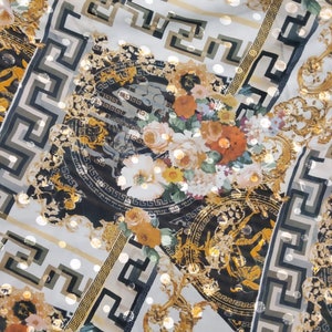 Op de afbeelding: Witte stof met een zwart en gouden patroon. Het patroon heeft een Grieks sleutelpatroon, bloemmotieven en een herhalend patroon van cirkels.