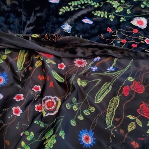 Op de afbeelding: Zwarte fluwelen stof met een borduurwerkontwerp met bloemen. Het borduurwerk heeft kleurrijke bloemen, bladeren en stelen op een zwarte achtergrond.
