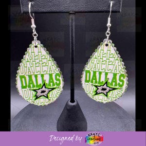 May include: A pair of teardrop-shaped earrings with a white background and green and black Dallas Stars logo. The earrings are decorated with rhinestones.