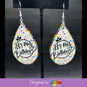 May include: Teardrop-shaped earrings with a white background, colorful confetti, and the text "It's my Birthday." The edges are lined with colorful rhinestones, and the earrings hang from silver hooks.