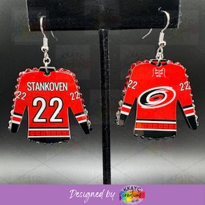 May include: A pair of red and black hockey jersey earrings, each featuring the number 22. One earring displays the name "STANKOVEN", the other the Carolina Hurricanes logo. Both have black and white striped sleeves and are embellished with red gems.
