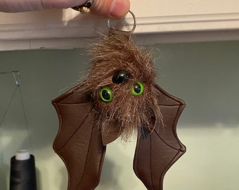 May include: A brown fuzzy bat keychain with green eyes and brown leather wings.