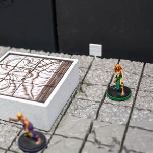 May include: A tabletop game setup featuring miniature figurines and a game board. The board is a white square with a brown and white design. Several figurines are placed around the board, including a horned creature and several human figures.