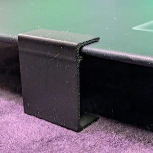 May include: A black metal bracket attached to the corner of a green surface. The bracket is rectangular and appears to be made of a sturdy material. The green surface has a smooth texture and is set against a purple background.