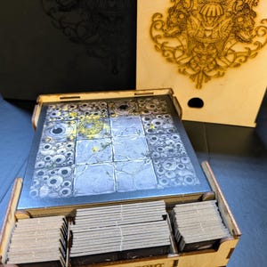 Puede incluir: Una caja de madera con una ilustración en blanco y negro de una criatura con múltiples ojos en la tapa. La caja está abierta y contiene un tablero de juego y varias barajas de cartas. La caja está etiquetada como "BLACK KNIGHT".