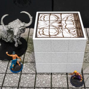 May include: A tabletop gaming scene featuring a gray cube-shaped terrain piece with a detailed top surface. Miniature figures of a monster and adventurers are positioned around the cube, suggesting a role-playing game setting.