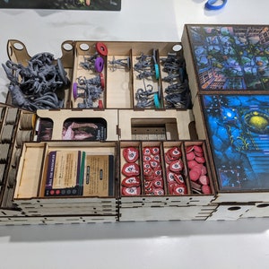 May include: A wooden organiser for a board game with multiple compartments. The organiser has a grey plastic monster, red and purple plastic figures, and a stack of cards with the text "Kill the Messenger".