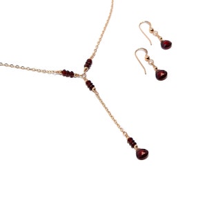 Puede incluir: Un conjunto de collar y pendientes a juego en tono dorado. El collar presenta un dise&ntilde;o en forma de Y con peque&ntilde;as cuentas de color rojo oscuro y un colgante m&aacute;s grande en forma de l&aacute;grima. Los pendientes tienen un dise&ntilde;o similar.
