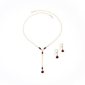 Puede incluir: Un conjunto de collar y pendientes de color dorado. El collar presenta un dise&ntilde;o en forma de Y con peque&ntilde;as cuentas de color rojo oscuro y una cuenta en forma de l&aacute;grima m&aacute;s grande en el extremo. Los pendientes tienen un dise&ntilde;o similar.