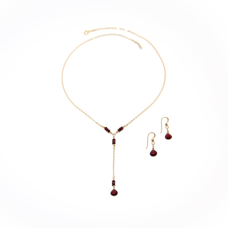 Puede incluir: Un conjunto de collar y pendientes de color dorado. El collar presenta un dise&ntilde;o en forma de Y con peque&ntilde;as cuentas de color rojo oscuro y una cuenta en forma de l&aacute;grima m&aacute;s grande en el extremo. Los pendientes tienen un dise&ntilde;o similar.