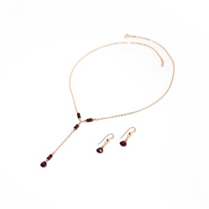 Puede incluir: Un conjunto de collar y pendientes de color dorado. El collar tiene un dise&ntilde;o en forma de Y con peque&ntilde;as cuentas de color rojo oscuro y una cuenta m&aacute;s grande en forma de l&aacute;grima en el extremo. Los pendientes presentan una sola cuenta en forma de l&aacute;grima.