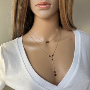 Puede incluir: Un collar dorado de varias capas con peque&ntilde;as cuentas de color rojo oscuro. La capa superior es una barra simple, mientras que la capa inferior tiene una ca&iacute;da vertical con una sola cuenta en el extremo. El collar se muestra en un maniqu&iacute; con una camiseta blanca con cuello en V.