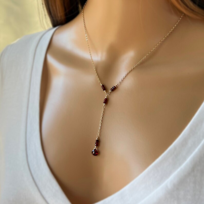 Puede incluir: Un delicado collar dorado con un dise&ntilde;o en forma de Y. El collar presenta peque&ntilde;as piedras preciosas redondas de color rojo intenso y una sola piedra preciosa en forma de l&aacute;grima al final de la ca&iacute;da. El collar se usa sobre una blusa blanca.