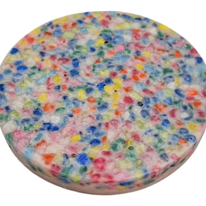 May include: A circular coaster with a speckled pattern. The coaster is made up of small, multi-coloured shapes, including blue, red, yellow, and pink, set against a pale pink base. The coaster is approximately 10 cm in diameter.