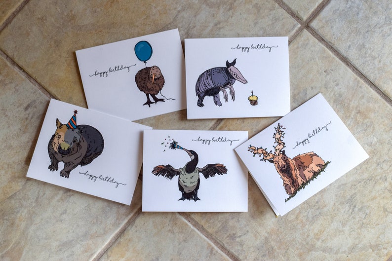 Pu&ograve; includere: Cinque diverse cartoline di compleanno con illustrazioni di animali. Ogni cartolina ha il testo "buon compleanno" scritto su di essa. Gli animali sono un wombat con un cappello da festa, un kiwi con un palloncino, un armadillo, un cormorano che suona un trombetta da festa e un cervo con le corna.