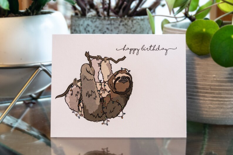 Pu&ograve; includere: Un biglietto d'auguri con un'illustrazione di un bradipo appeso a un ramo con luci di Natale avvolte intorno a lui. Il biglietto ha il testo "buon compleanno" scritto su di esso.