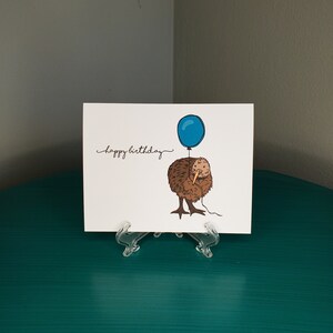 Pu&ograve; includere: Un biglietto d'auguri con un kiwi marrone che tiene un palloncino blu. Il biglietto dice "buon compleanno" in corsivo.