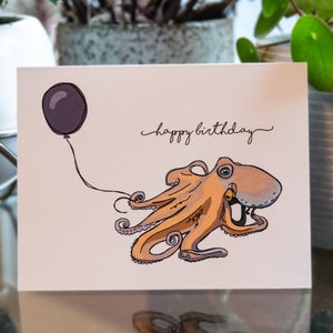 Pu&ograve; includere: Un biglietto d'auguri con un polpo dei cartoni animati che tiene un palloncino viola. Il biglietto dice "buon compleanno" in corsivo.