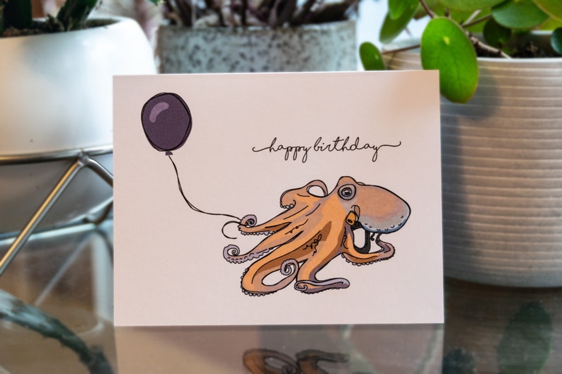 Pu&ograve; includere: Un biglietto d'auguri con un polpo dei cartoni animati che tiene un palloncino viola. Il biglietto dice "buon compleanno" in corsivo.