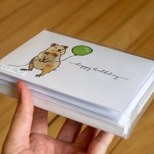 Pu&ograve; includere: Un multipack di biglietti d'auguri con un'illustrazione di un quokka che tiene un palloncino verde. I biglietti sono bianchi con il testo "buon compleanno" scritto in corsivo nero.