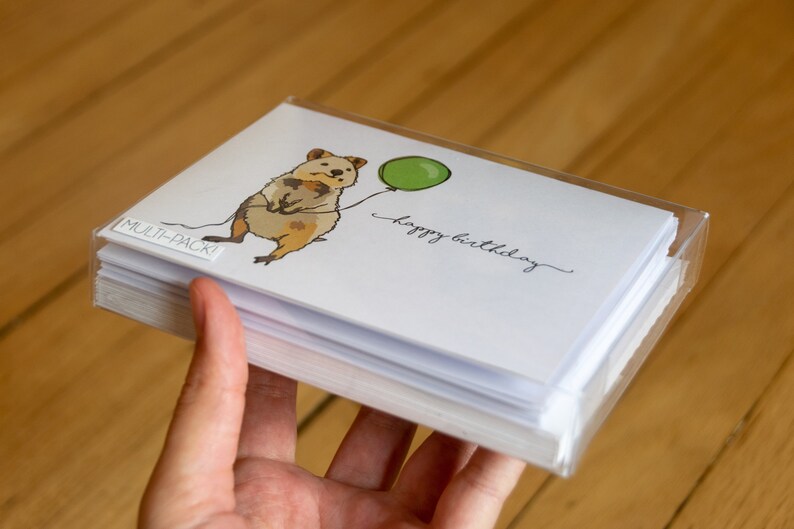 Pu&ograve; includere: Un multipack di biglietti d'auguri con un'illustrazione di un quokka che tiene un palloncino verde. I biglietti sono bianchi con il testo "buon compleanno" scritto in corsivo nero.