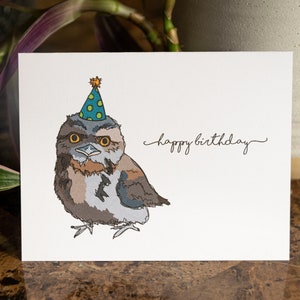 Pu&ograve; includere: Un biglietto di auguri di compleanno con un'illustrazione di un gufo marrone e grigio che indossa un cappello da festa verde e blu con stelle gialle. Il testo "happy birthday" &egrave; scritto in corsivo sulla carta.