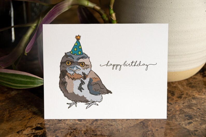 Pu&ograve; includere: Un biglietto di auguri di compleanno con un'illustrazione di un gufo marrone e grigio che indossa un cappello da festa verde e blu con stelle gialle. Il testo "happy birthday" &egrave; scritto in corsivo sulla carta.