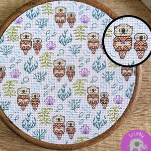 May include: A circular wooden embroidery hoop containing white fabric with a cross-stitch pattern of brown sea otters, green seaweed, purple seashells, and small blue fish. A circular inset shows a close-up of one otter pair.