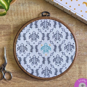 Puede incluir: Un aro de bordado blanco con un diseño de tortuga geométrica en negro y turquesa. El aro está rodeado por un marco de madera.