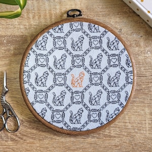 Puede incluir: Un aro de bordado blanco con un patrón de gato geométrico en blanco y negro. Un gato es naranja en el centro del aro.