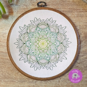 May include: A cross-stitch mandala, featuring a detailed design in green hues, displayed on white fabric within a round wooden embroidery hoop. The design showcases intricate patterns and gradients, creating a visually appealing piece of art.