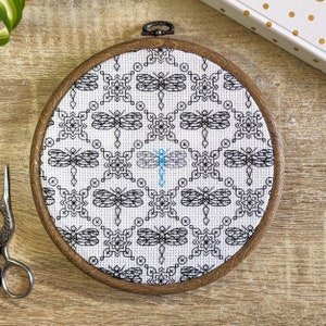 May include: A white embroidery hoop with a black and white geometric pattern featuring dragonflies. One dragonfly is blue.