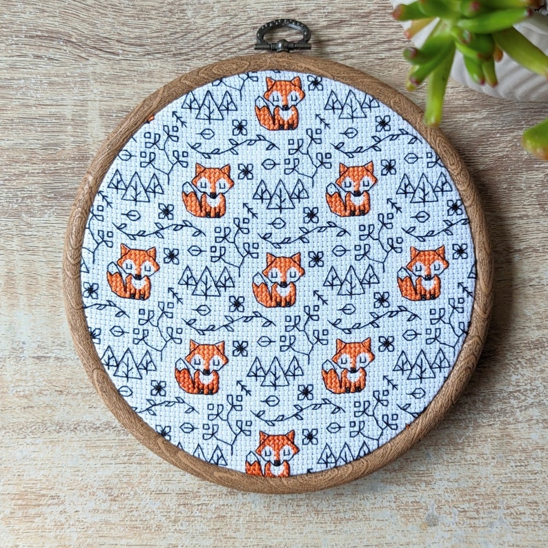May include: A white embroidery hoop with a brown wooden frame. The fabric inside the hoop features a repeating pattern of orange foxes and black trees on a white background.