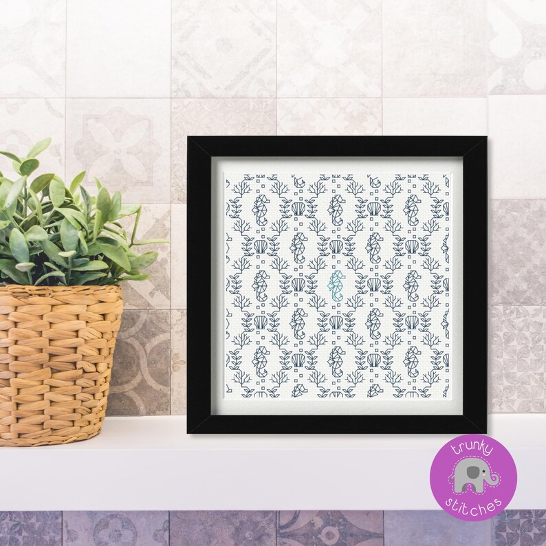 May include: A framed artwork featuring a repeating pattern of seahorses, coral, and shells in navy blue on a white background. The artwork is in a black frame and sits on a white shelf. A potted plant in a woven basket is to the left.