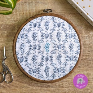 Op de afbeelding: Een ronde houten borduurring met een kruissteekpatroon van zeepaardjes, schelpen en zeewier in blauwtinten op een witte achtergrond. De ring ligt op een houten oppervlak, met een zilveren schaar. Het ontwerp heeft een nautisch thema.