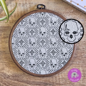 May include: A circular wooden embroidery hoop with a cross-stitch design of skulls and crossbones in black thread on white fabric. A magnified view highlights the stitching. The hoop has a metal hanging loop.
