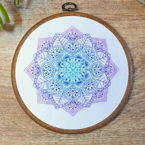 May include: A colourful mandala embroidery pattern in shades of blue, purple and pink on a white canvas, framed in a wooden hoop.