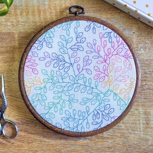 Peut inclure: Un cercle de broderie circulaire avec un fond en tissu blanc et un motif géométrique coloré de feuilles. Le motif est dans des tons de rose, bleu, vert et jaune.