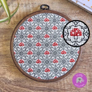 May include: A round, wooden embroidery hoop featuring a cross-stitch design. The design includes a repeating pattern of red mushrooms on a white and black geometric background. A close-up shows the detailed stitching.