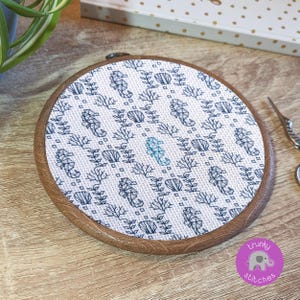 May include: A round embroidery hoop featuring a cross-stitch design of seahorses, seashells, and coral in shades of blue on a white background. The wooden hoop is approximately 6 inches in diameter. The logo "trunky stitches" is visible in the lower right corner.