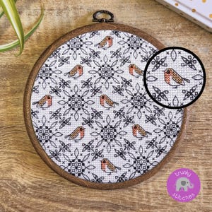 Op de afbeelding: Een ronde houten borduurring met een kruissteekontwerp. Het ontwerp bevat een herhalend patroon van kleine vogels met rode borsten en zwart-witte sneeuwvlokachtige vormen. De ring heeft een diameter van ongeveer 15 cm.
