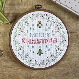 Peut inclure: Un cerceau de broderie blanc avec une couronne de verdure, des baies rouges et le texte "Merry Christmas" en noir et rouge. Un sapin de Noël vert avec une étoile est au centre.