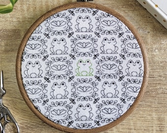 May include: A circular wooden embroidery hoop containing white fabric with a repeating cross-stitch pattern of black frogs and floral motifs. One central frog is stitched in light green thread, while all other frogs are stitched in black thread.