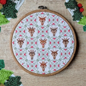 May include: A white embroidery hoop with a cross-stitch design of a repeating pattern of stylized reindeer heads and red and green floral motifs.