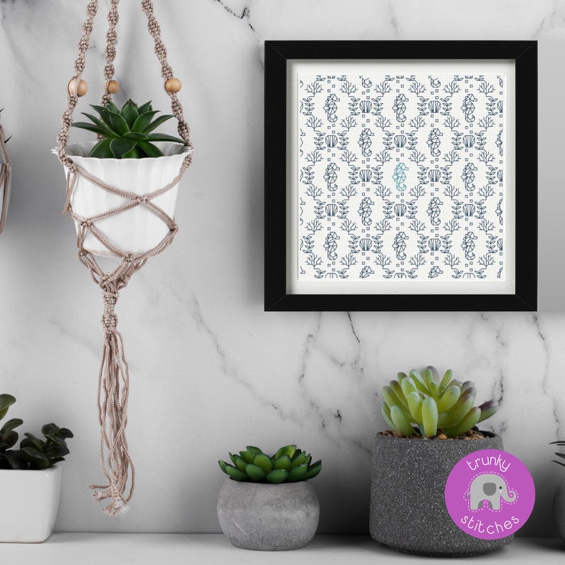 May include: A white ceramic pot with a green succulent plant hangs in a macrame plant hanger. A framed print with a blue and white seahorse pattern is on the wall. Other potted succulents are on a white surface.