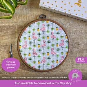 May include: A round wooden embroidery hoop with a flamingo blackwork pattern. The pattern features pink flamingos, green palm leaves, and yellow pineapples on a white background. A pair of silver scissors and a purple label with text are visible.
