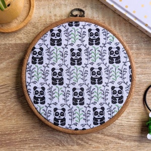 Puede incluir: Un aro de bordado redondo con un fondo de tela blanca que presenta un patrón repetido de osos panda negros y blancos y hojas verdes.