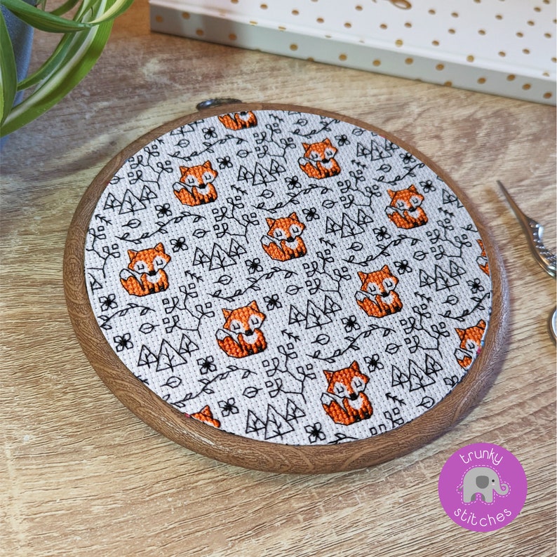 May include: A white embroidery hoop with a brown wooden frame, featuring a repeating pattern of orange foxes, black trees, and leaves on a white background. The hoop is on a wooden surface.