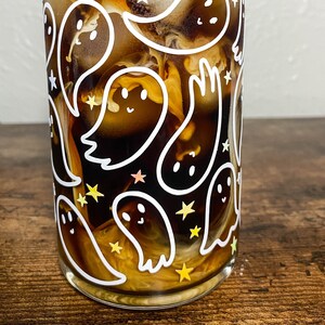 May include: A glass with a white ghost and star pattern filled with iced coffee. The coffee is a dark brown color and the milk is a light brown color.