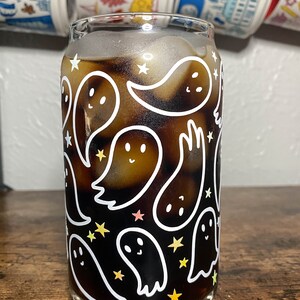 May include: A glass with a black and white ghost pattern and a dark brown liquid inside. The glass has a white rim and a clear base. The ghosts are outlined in white and have small eyes and smiles. There are also small white stars scattered around the ghosts.